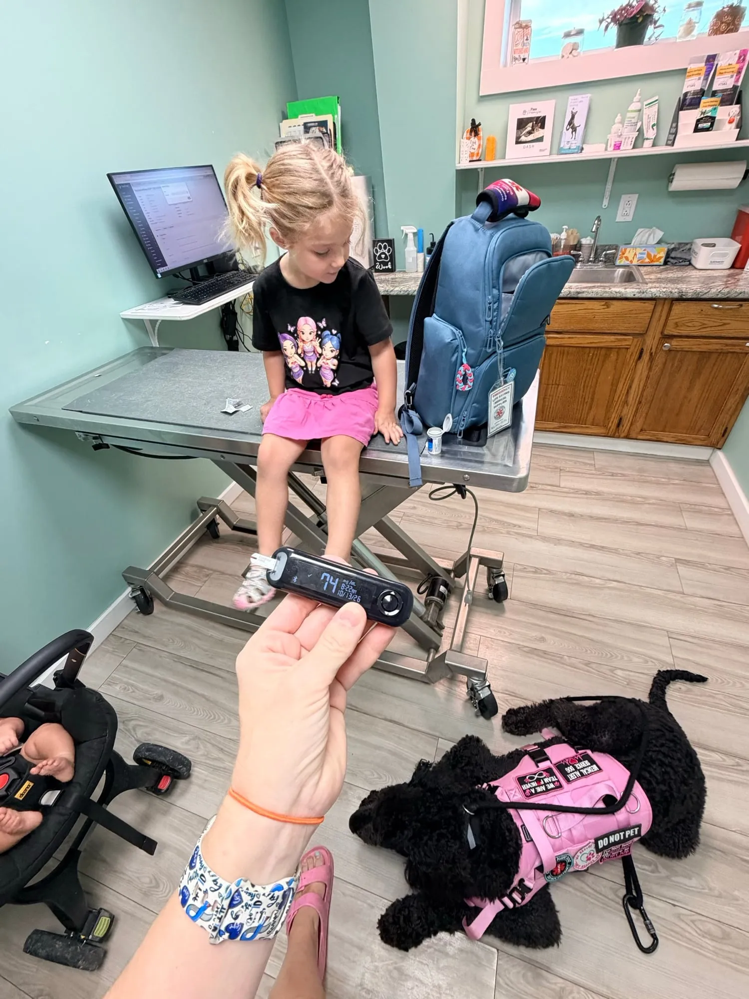 A little girl perched on a grooming table, smiling, with a dog in a pink service vest nearby
